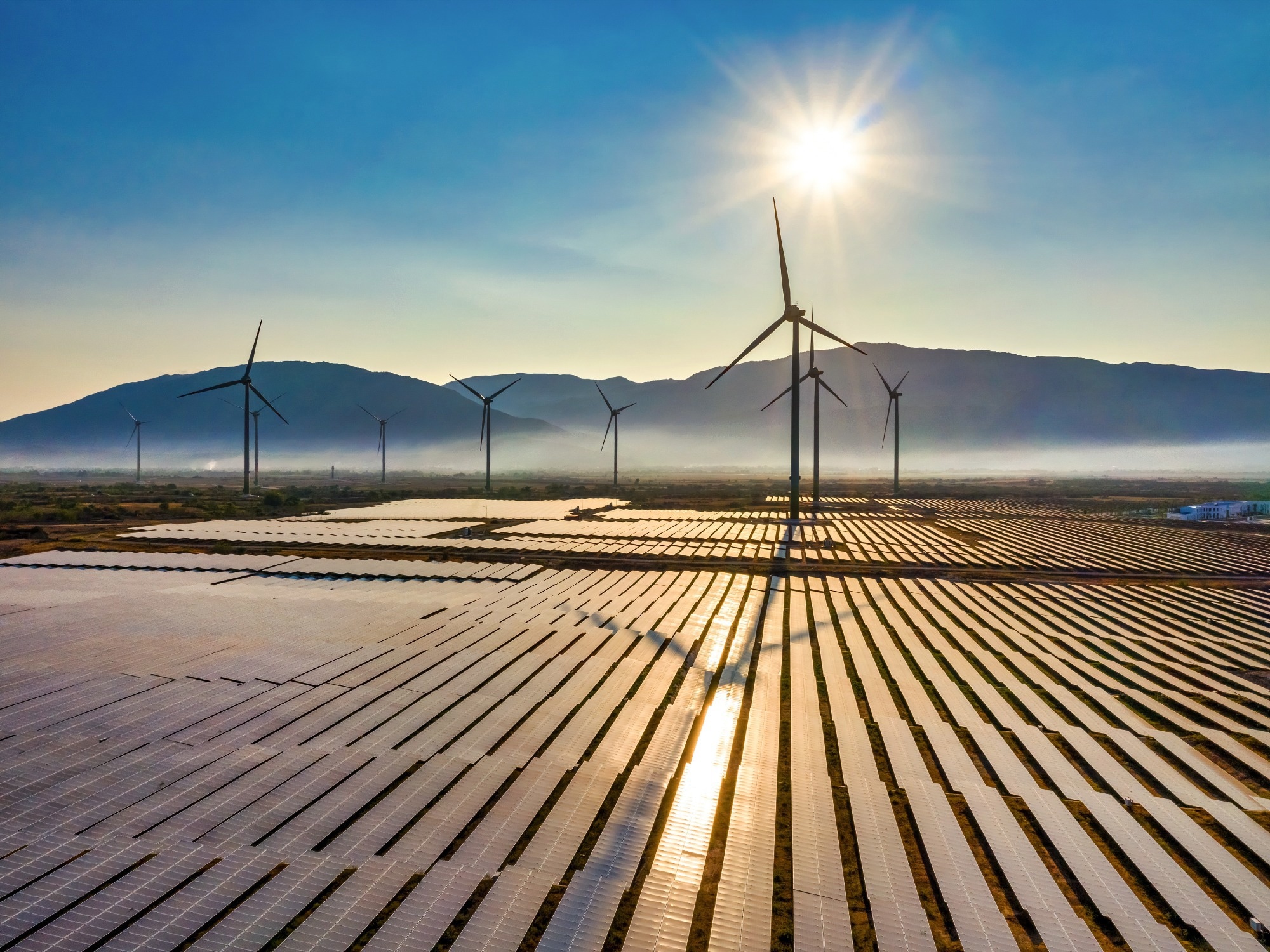 A vast landscape showing a combination of solar panels and wind turbines under a clear sky, with mountains in the background.