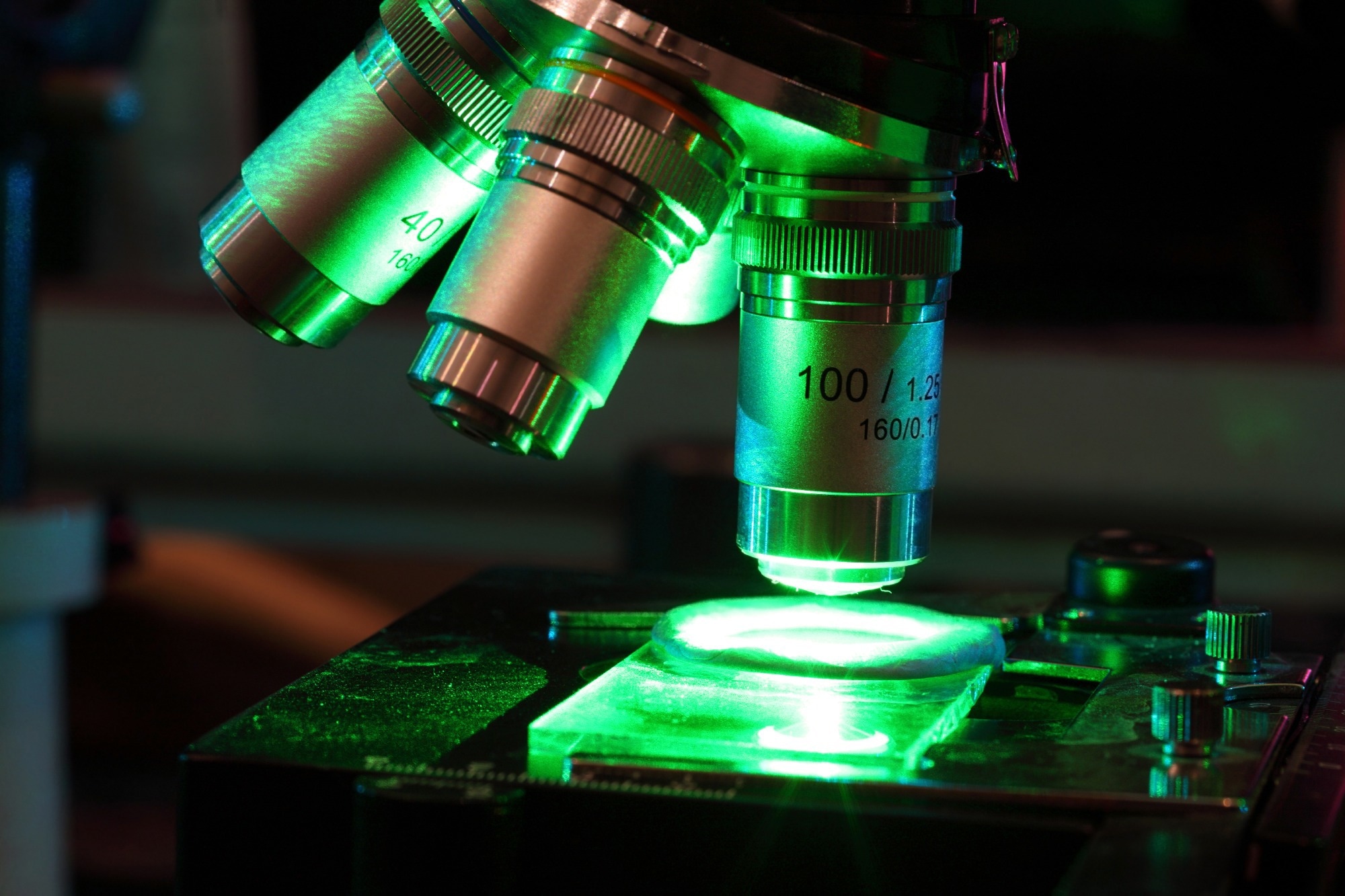 lose-up view of a microscope illuminated with green light, focusing on the lens assembly and a glass slide on the stage.