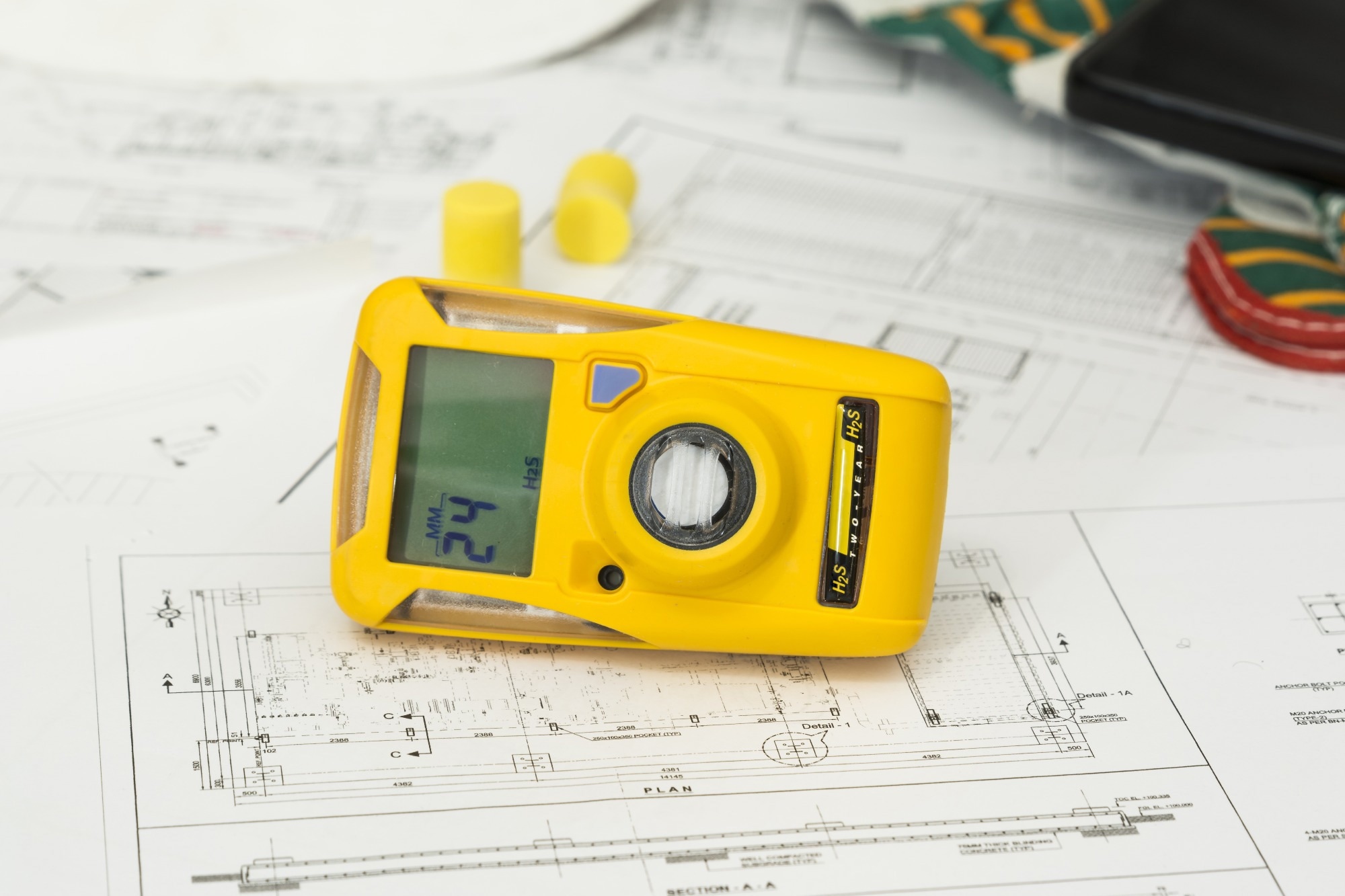 A portable yellow gas detector placed on top of engineering blueprints, displaying a gas concentration reading on its screen. The device is used for detecting hazardous gases in industrial and environmental settings.