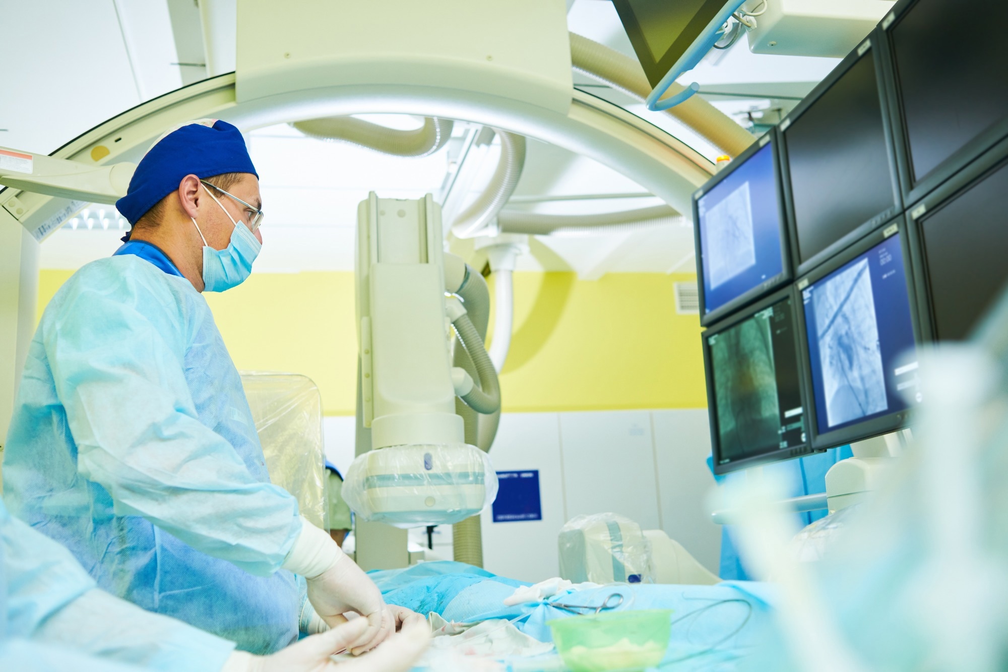 A doctor in scrubs and a face mask performing surgery, with imaging screens.